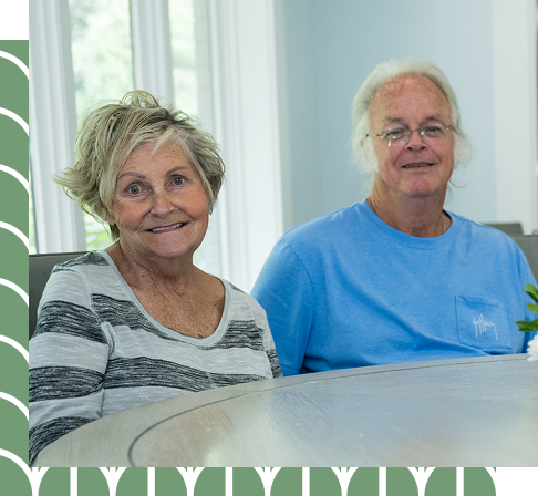 An elderly couple seated at a table—he in a blue t-shirt, she in a green and white striped shirt—smiling together, reflecting the milestones and memories that shape the SECU Caring House story.