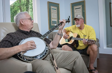 Two elderly gentlemen enjoying a moment of warmth and dignity in the communal living area of SECU Caring House, playing music together—one on the banjo, the other on the guitar—reflecting the home’s spirit of care and com
